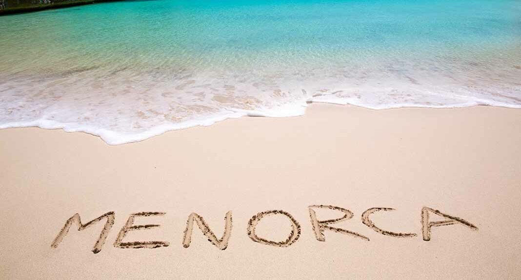 Texto "MENORCA" escrito en la arena de una playa, con agua turquesa de fondo.