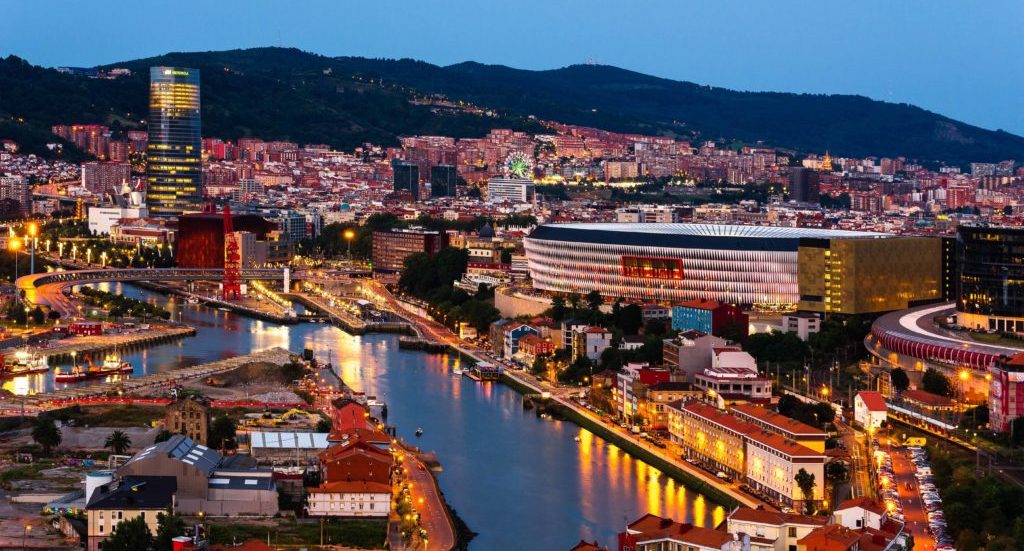 Vista panorámica de una ciudad con un río, montañas y un estadio iluminado en Bilbao.